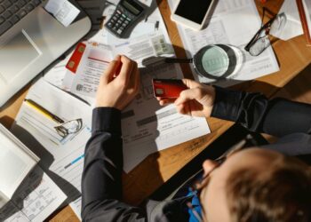 This image depicts a typical overwhelmed worker surrounded by an array of financial documents, electronic devices, and various office materials spread across a wooden desk. In the midst of chaos, the individual appears focused, with hands busy sorting and analyzing content. This scene captures the essence of contemporary work stress, emphasizing the pressures and clutter often associated with demanding office jobs. Sunlight filtering through hints at the passage of time, possibly suggesting a long day spent at work.