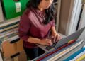 In the warmth of a sunlit room, a woman deeply engaged in her work peers intently at her laptop screen, surrounded by neatly organized files and office supplies. The orderliness of her workspace, with color-coded folders and clearly labeled storage, suggests a meticulous and systematic approach to her tasks at hand. The focused atmosphere and the soft daylight streaming through the window create an environment conducive to productivity and concentration.