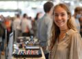 A cheerful young woman stands at the forefront of a technology expo, her eyes sparkling with curiosity and excitement. Surrounded by a crowd of fellow enthusiasts, she is captured exploring a range of advanced electronic devices laid out before her. Her focused attention towards the equipment showcases her interest in the latest technological advancements. This vibrant setting, filled with eager participants and cutting-edge technology, highlights the dynamic nature of tech expos and their role in fostering innovation and engagement among tech aficionados.